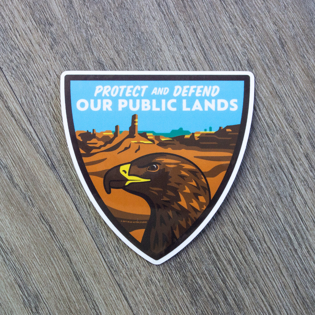 A shield shaped vinyl sticker with an eagle and desert scene and the words Protect And Defend Our Public Lands at the top.