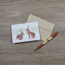 Load image into Gallery viewer, A note card featuring an illustration of two rabbits sitting on a natural brown envelope with a pen next to them.
