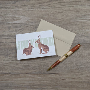 A note card featuring an illustration of two rabbits sitting on a natural brown envelope with a pen next to them.