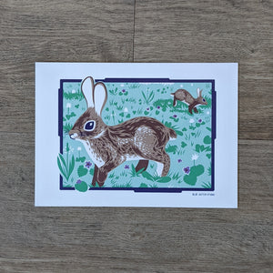 An art print of an illustration of two rabbits in a grassy field that includes purple and white flowers.