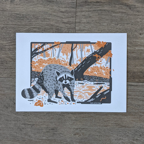 An art print of a raccoon by a creek surrounded by red and orange fall leaves.
