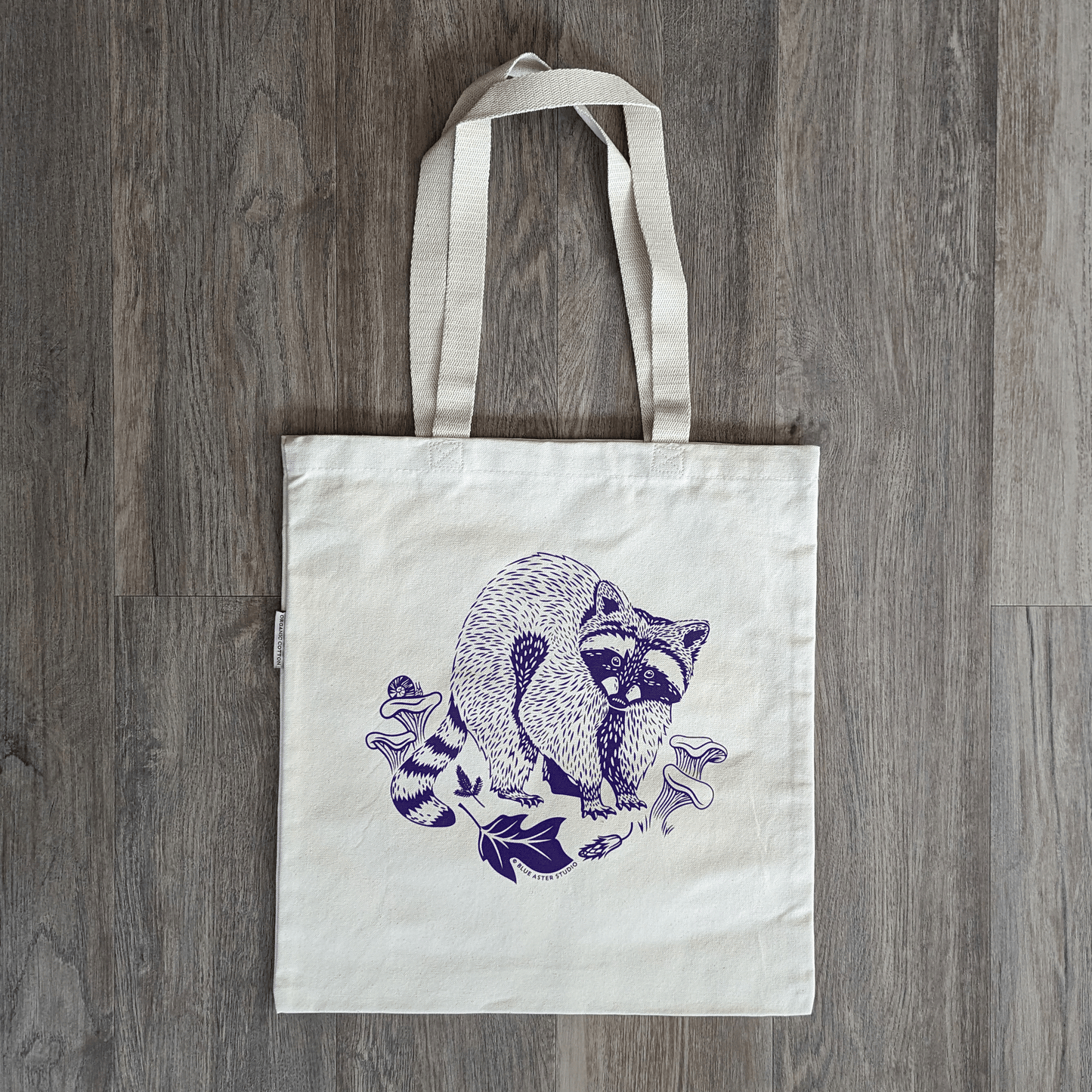 An organic cotton canvas tote bag with an illustrated design of a racoon surrounded by edible chanterelle mushrooms and leaves of a tulip poplar tree. A snail perches on one of the mushrooms.