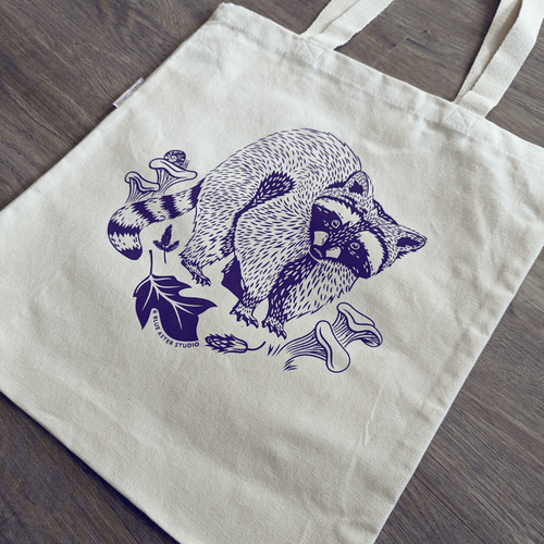 An organic cotton canvas tote bag with an illustrated design of a racoon surrounded by edible chanterelle mushrooms and leaves of a tulip poplar tree. A snail perches on one of the mushrooms.