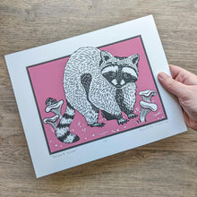 Load image into Gallery viewer, The 8x10 raccoon and shrooms screen print with the pink background being held in a hand to show scale.