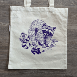 An organic cotton canvas tote bag with an illustrated design of a racoon surrounded by edible chanterelle mushrooms and leaves of a tulip poplar tree. A snail perches on one of the mushrooms.