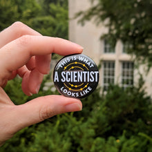 Load image into Gallery viewer, The black This Is What A Scientist Looks Like button being held up in front of limestone campus buildings and greenery.