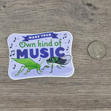 Load image into Gallery viewer, A vinyl sticker with a katydid and cicada illustration on it with the words &quot;Make Your Own Kind Of Music&quot; sitting next to a USD quarter for scale.