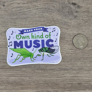 A vinyl sticker with a katydid and cicada illustration on it with the words "Make Your Own Kind Of Music" sitting next to a USD quarter for scale.