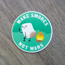 Load image into Gallery viewer, A round vinyl sticker with an illustration of a cute marshmallow toasting its butt on a campfire with the words Make S’Mores Not Wars.