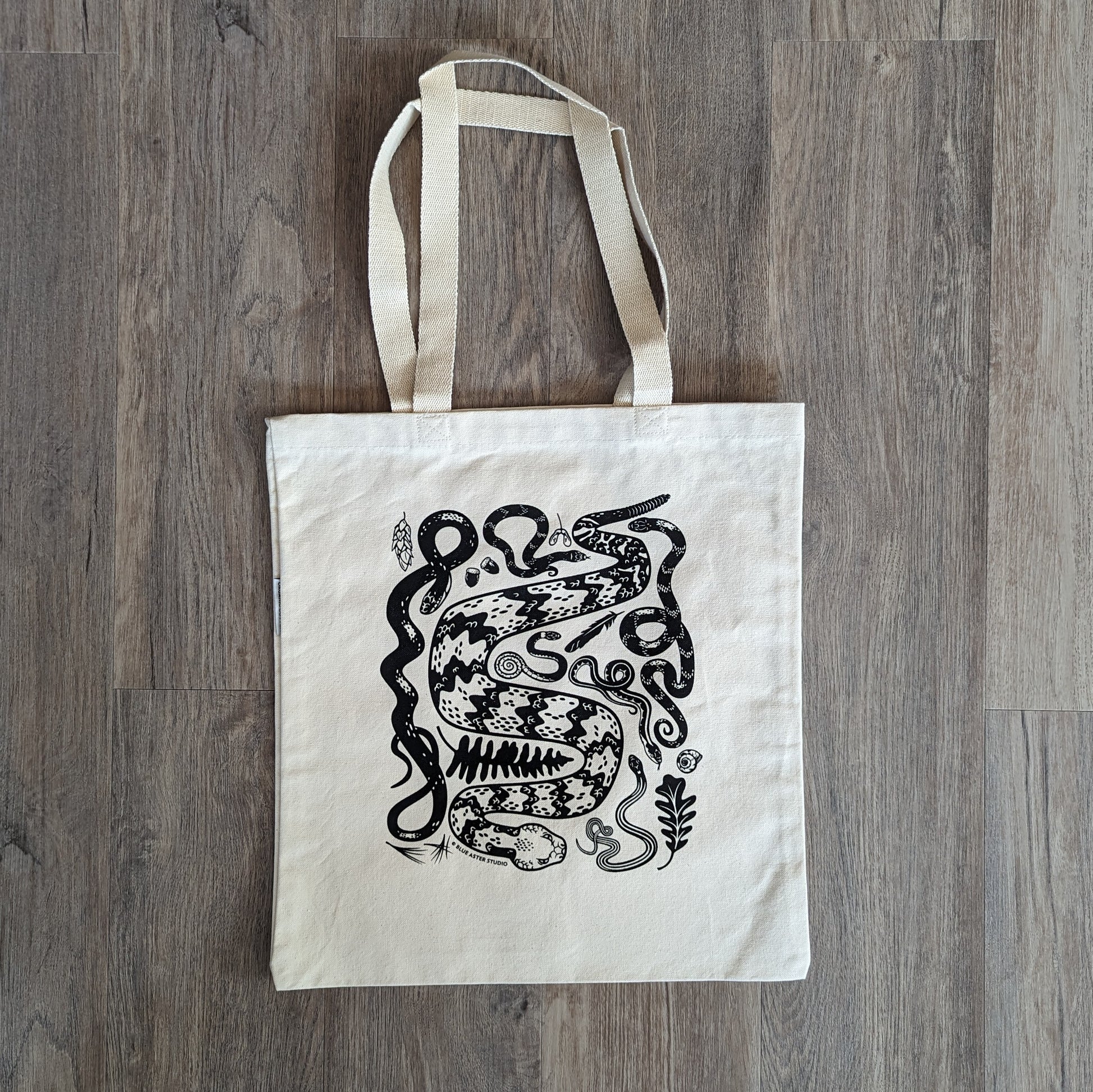 A sturdy canvas tote bag with woven handles featuring an illustration of a collection of seven snakes native to the eastern United States. The snakes include a timber rattlesnake, black rat snake, eastern hognose, ringneck, water snake, garter snake, and ribbon snake.