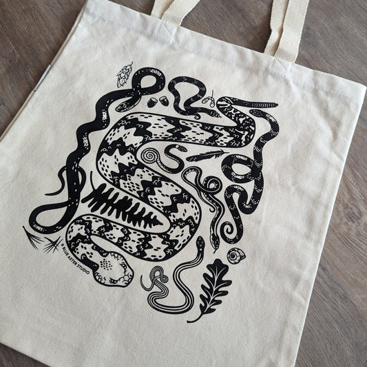 A sturdy canvas tote bag with a collection of illustrated snakes screen printed on it in black ink. The snakes include a timber rattlesnake, black rat snake, eastern hognose, ringneck, water snake, garter snake, and ribbon snake.