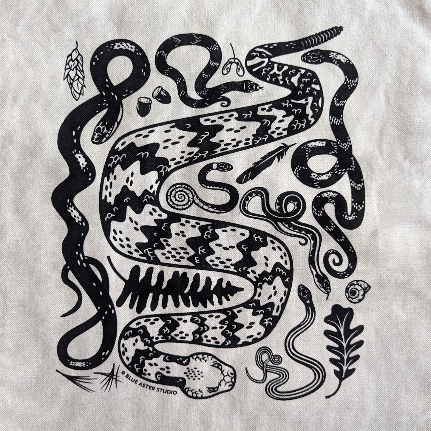 A black design of snakes and natural objects you might find on a forest floor. The snakes include a timber rattlesnake, black rat snake, eastern hognose, ringneck, water snake, garter snake, and ribbon snake.