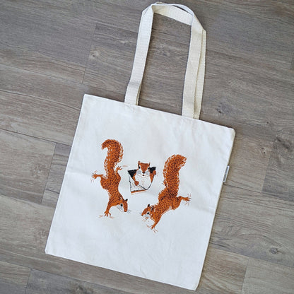 A squirrel tote bag featuring three illustrations of squirrels climbing and peeking out of the tote. The bag is made of a durable organic cotten fabric.