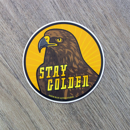 A round vinyl sticker with an illustration of a golden eagle surrounded by a yellow sunburst pattern and the words Stay Golden.