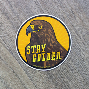 A round vinyl sticker with an illustration of a golden eagle surrounded by a yellow sunburst pattern and the words Stay Golden.