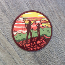 Load image into Gallery viewer, A round vinyl sticker with the silhouettes of two hikers in the red rocks desert and the words Take A Hike at the bottom.