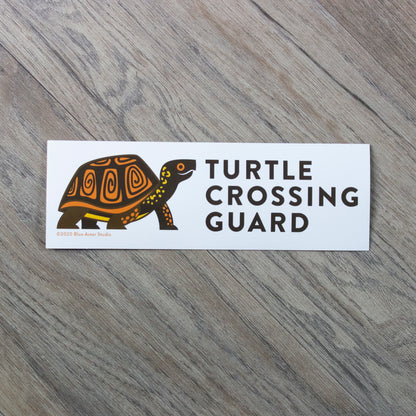 A 2.5 by 7.5 inch white vinyl bumper sticker with an illustration of a box turtle and the words "Turtle Crossing Guard" to the right of the illustration.