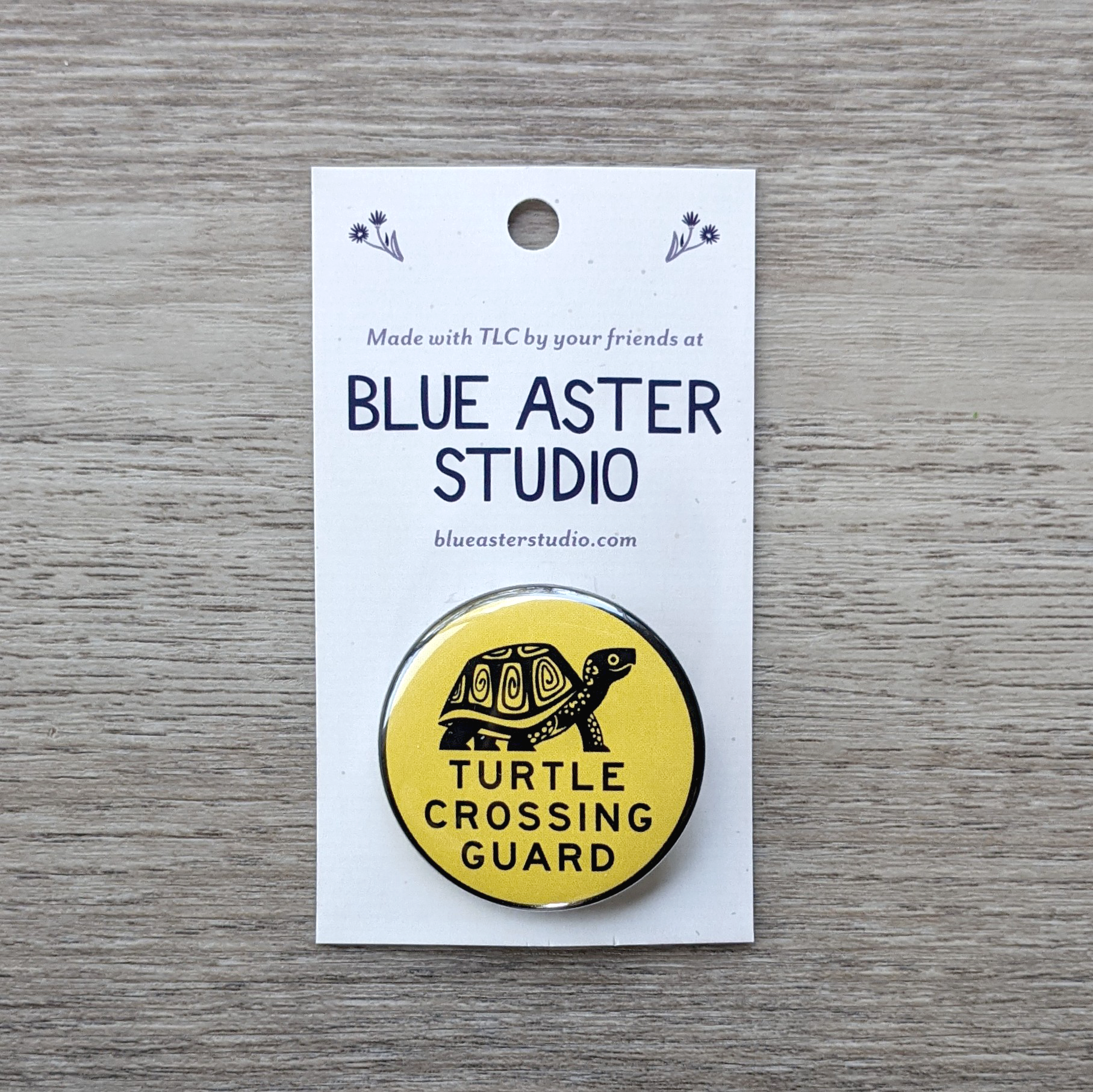 A 1.5 inch pinback button on a backer card. The button has an illustration of a box turtle and the words "Turtle Crossing Guard" in black on a yellow background.