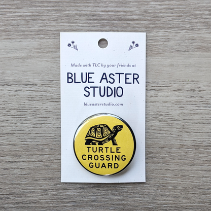 A 1.5 inch pinback button on a backer card. The button has an illustration of a box turtle and the words "Turtle Crossing Guard" in black on a yellow background.