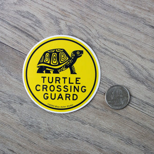 A 3 inch round vinyl sticker with an illustration of a turtle and the words " Turtle Crossing Guard" next to a USD quarter for scale.