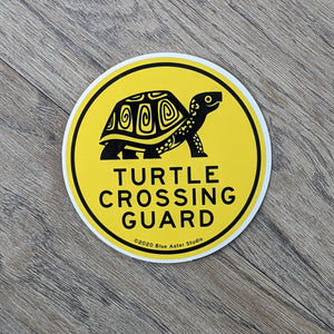 A 3 inch round vinyl sticker with the words "Turtle Crossing Guard" and an illustration of a box turtle in black on a yellow background.