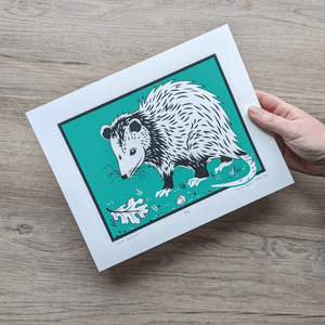 A hand holds a screen printed art on 8x10 paper. An opossum stands against a teal background with an oak leaf and acorn lying near it.